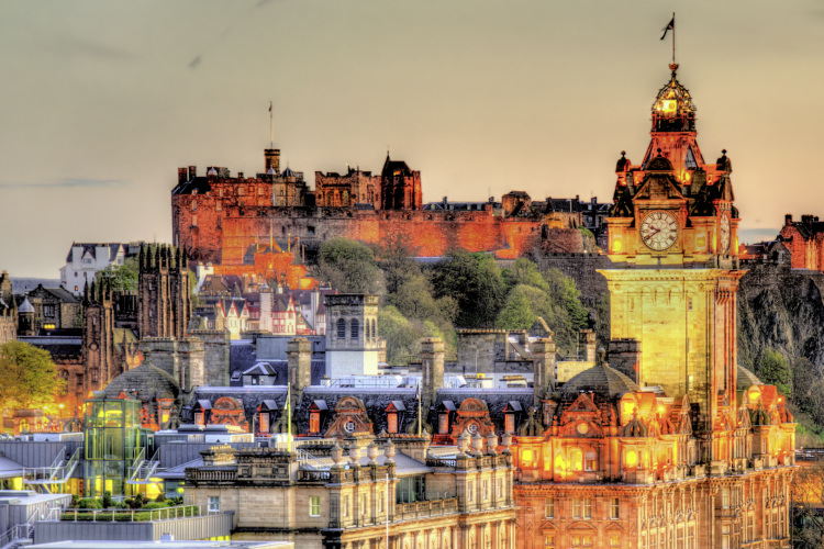 Stimmungsvolle Stadtansicht von Edinburgh bei Dämmerung mit dem markanten Balmoral Hotel Uhrturm im Vordergrund und dem beleuchteten Castle dahinter.