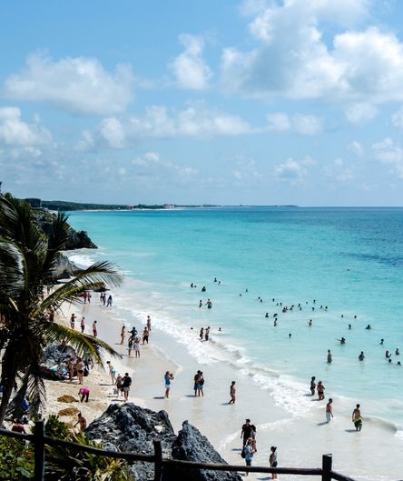 Ein belebter, weißer Sandstrand mit türkisfarbenem Meer. Viele Menschen schwimmen im Wasser oder spazieren am Ufer, eingerahmt von Felsen und Palmen. Ein belebter, weißer Sandstrand mit türkisfarbenem Meer. Viele Menschen schwimmen im Wasser oder spazieren am Ufer, eingerahmt von Felsen und Palmen.
